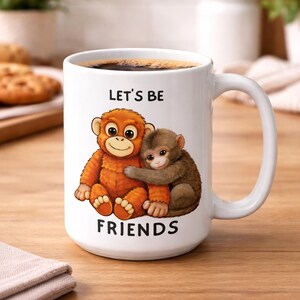 Könnte beinhalten: Weiße Keramik-Tasse mit Henkel, gefüllt mit Kaffee. Die Tasse zeigt eine Cartoon-Illustration von zwei sich umarmenden Affen mit dem Text "LET'S BE FRIENDS" auf der Vorderseite. Die Affen sind orange und braun.