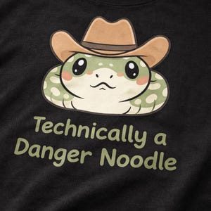 Puede incluir: Camiseta negra con una serpiente de dibujos animados con sombrero de vaquero. La serpiente es verde y blanca con mejillas rosas. El texto en la camiseta dice "Technically a Danger Noodle" en verde.