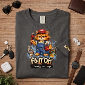 Może przedstawiać: Ciemnoszara koszulka z kreskówkowym kotem hydraulikiem z kluczem, w kombinezonie i czerwonej czapce. Koszulka ma napis "Fluff Off" i "I don't give a crap". Humorystyczny design.