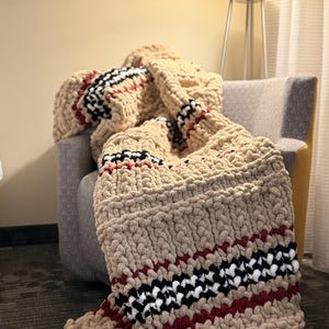May include: A chunky knit blanket in a neutral beige color, draped over a gray armchair. The blanket features a bold diagonal pattern of black, white, and red stripes, adding a striking contrast to the solid beige. The thick, textured yarn suggests a cozy and warm feel.
