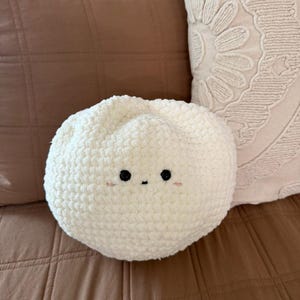 May include: A round, off-white crochet pillow with black eyes and a small mouth. The pillow is placed on a brown quilted surface, with a brown and cream-colored pillow in the background.