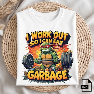 Op de afbeelding: Wit t-shirt met een cartoon schildpad die gewichten heft, met de tekst "I WORK OUT SO I CAN EAT GARBAGE". De schildpad is groen met een rode hoofdband. Het ontwerp heeft oranje en gele accenten.