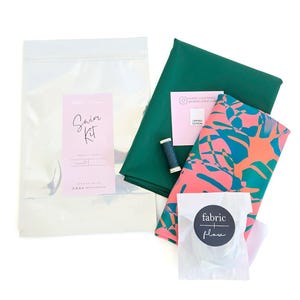 May include: A sewing kit for swimwear, featuring fabric, thread, and instructions. The kit includes a clear plastic bag with a pink label that reads "Swim Kit". There is also green fabric, patterned fabric, and a spool of blue thread.