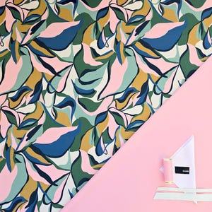 May include: A colorful fabric with a leaf pattern in shades of pink, blue, green, and gold. The fabric is diagonally placed on a pink background, with a spool of pink thread, a white fabric triangle, and a label that says "DAMN".