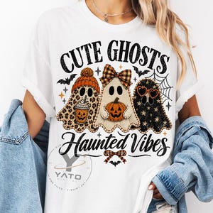 May include: A white t-shirt featuring a graphic of three ghosts with the text CUTE GHOSTS HAUNTED VIBES. The first ghost has a leopard print pattern and orange beanie, the second a plaid bow and pumpkin, and the third a black star pattern and heart glasses.