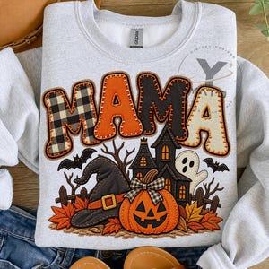 Op de afbeelding: Een lichtgrijs sweatshirt met ronde hals en het woord MAMA in grote, gestructureerde letters met ruitpatronen, oranje, zwart en crème. Daaronder staat een afbeelding van een heksenhoed, een pompoen, een spookhuis en een geest.
