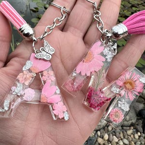 May include: Three resin keychains with the letters "A", "M", and "N" embedded with pink flowers and silver flakes. Each keychain has a silver chain, a pink tassel, and a decorative charm.