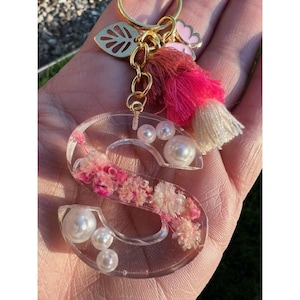 Op de afbeelding: Een sleutelhanger met de letter "S" van heldere hars met ingebedde roze bloemen en imitatieparels. De sleutelhanger heeft een goudkleurige metalen ketting, een kwast met roze en crèmekleurige draden en een bladbedel.