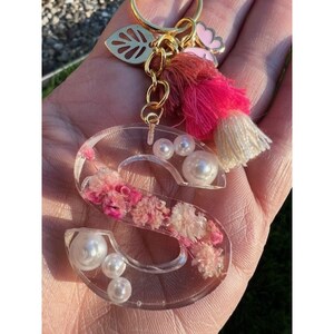 May include: A clear resin letter "S" keychain with embedded pink flowers and faux pearls. The keychain has a gold-colored metal chain, a tassel with pink and cream threads, and a leaf charm.