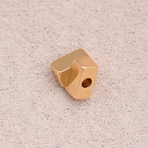 May include: A small, gold-colored geometric bead with a hexagonal shape and a single hole. The bead has a polished finish and is likely used for jewelry making or crafting. The design is modern and minimalist.