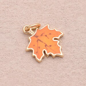 May include: A small maple leaf charm featuring a bright orange enamel fill set within a gold-toned metal frame. The leaf design includes detailed vein markings and a small circular jump ring attached at the top for easy connection to jewellery.