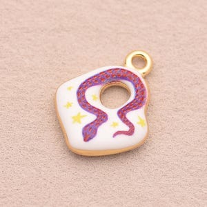 Puede incluir: Un dije de metal dorado con base de esmalte blanco, que presenta una serpiente texturizada en color morado y rojo enroscada alrededor de un recorte circular central, decorado con pequeñas estrellas amarillas. Incluye una argolla dorada.