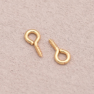 May include: Two small, gold-colored screw eye hooks. Each hook has a looped eye and a threaded screw end. The hooks are shown against a neutral, textured background, ideal for jewelry making or craft projects.