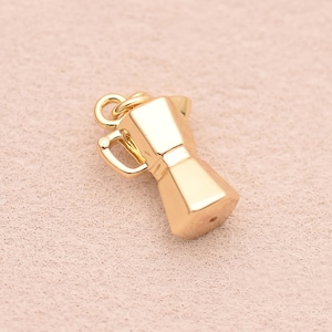 May include: A small, gold-toned metal charm shaped like a moka pot coffee maker. The charm features a faceted, geometric body with a handle on one side and a small circular loop at the top for attachment, showcasing a polished, reflective surface.