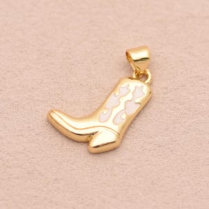 Op de afbeelding: Een goudkleurige metalen hanger in de vorm van een cowboylaars, met een gladde, gepolijste afwerking en een lichtroze emaille inlegdetail op de schacht van de laars. De hanger bevat een kleine, geïntegreerde goudkleurige ring aan de bovenkant.