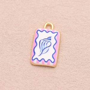 Könnte beinhalten: Ein rechteckiger goldfarbener Metallanhänger mit weißer Mitte und einer blauen Muschelabbildung. Der Anhänger ist von einer gewellten blauen Linie sowie vertikalen rosa und weißen Streifen umrandet und besitzt eine kleine goldene Öse oben.