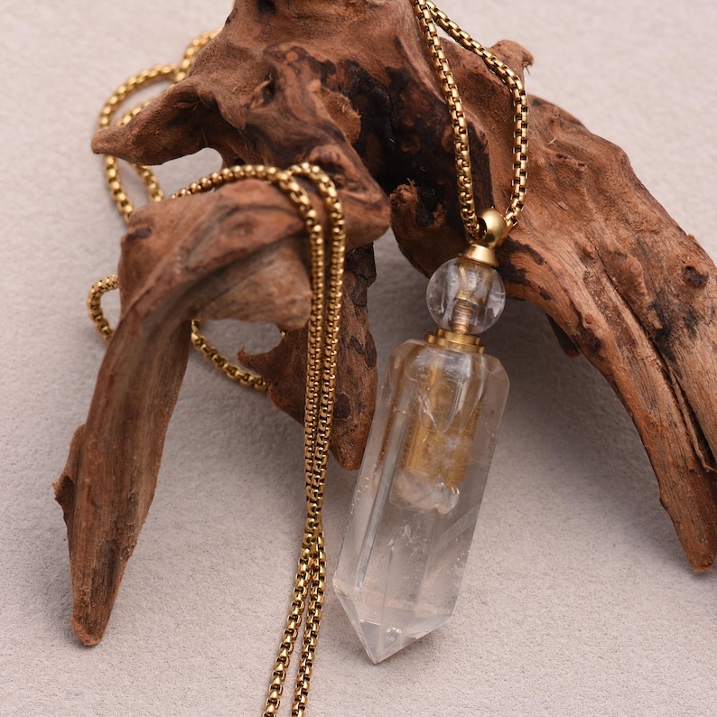 May include: A clear crystal pendant necklace with a gold-colored chain. The pendant is a clear, rectangular prism with a pointed bottom and a gold-colored cap. The chain is a delicate, gold-colored box chain. The necklace is displayed on a piece of driftwood.