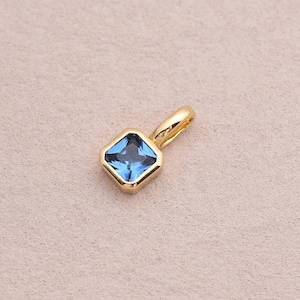 May include: A gold pendant with a square, blue gemstone. The gemstone is set in a gold bezel and has a small loop for a chain. The pendant is displayed on a neutral background.