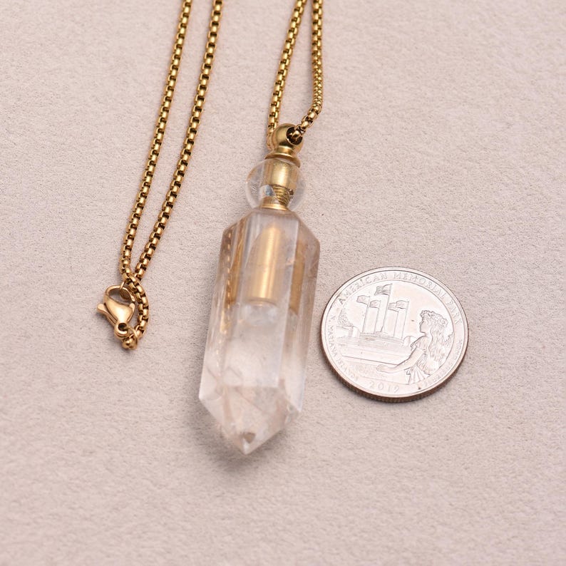 May include: A clear, faceted crystal pendant with a gold-colored metal cap and chain. The pendant is next to a U.S. quarter coin for size comparison. The chain has a lobster clasp.