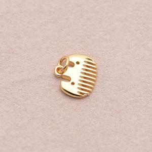 May include: A gold-coloured comb pendant with a small loop for attaching to a necklace or bracelet. The comb has a rounded shape with evenly spaced teeth and two small holes near the top. The pendant is made of shiny metal.