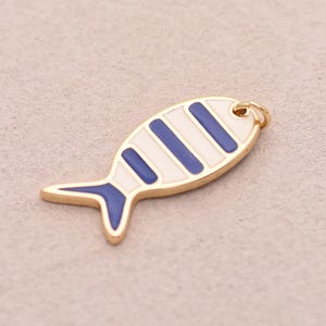 May include: A gold-toned metal fish charm featuring white and dark blue enamel stripes across the body and a solid dark blue tail. The fish-shaped pendant includes a small circular jump ring attached to the top for hanging or jewellery attachment.