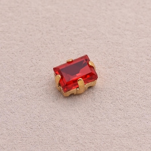 Puede incluir: Una gema roja rectangular y facetada engastada en un marco de metal dorado. La gema tiene un color intenso y rico y está montada con cuatro garras. El engaste tiene un diseño sencillo y elegante, adecuado para la fabricación de joyas.