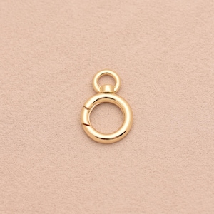 May include: A gold-toned circular spring ring clasp featuring a smooth, polished finish. The jewelry component includes a small, fixed loop at the top for attachment and a spring-loaded mechanism that allows the main ring to open and close securely.
