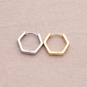May include: Two hexagonal hoop earrings, one silver-colored and one gold-colored, are displayed against a neutral background. The earrings have a geometric design and a hinged closure.