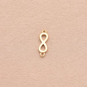 Puede incluir: Un conector de metal dorado con forma de símbolo de infinito en orientación vertical. La pieza tiene una superficie lisa y pulida con un pequeño bucle circular en los extremos superior e inferior, diseñado para sujetarse a cadenas o cordones.