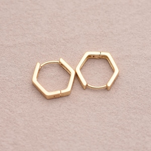 May include: A pair of gold-colored, geometric hoop earrings. Each earring features a hexagonal shape with a hinged closure. The earrings are displayed on a light pink surface.