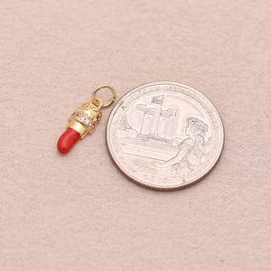 May include: A gold-toned lipstick charm featuring a bright red tip and a band of small clear stones. The charm includes a circular jump ring at the top for attachment. It is positioned next to a United States quarter coin for scale reference.