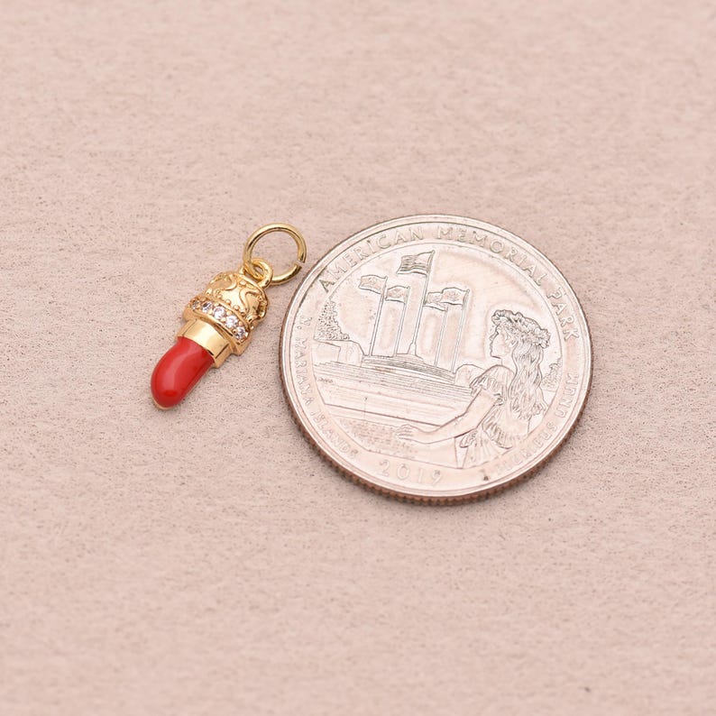 May include: A gold-toned lipstick charm featuring a bright red tip and a band of small clear stones. The charm includes a circular jump ring at the top for attachment. It is positioned next to a United States quarter coin for scale reference.