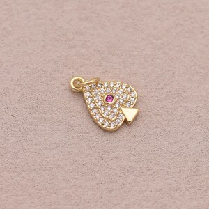 May include: A gold-toned spade-shaped charm featuring a pavé setting of clear white crystals and a single round pink stone centered in the middle. The charm includes a small circular jump ring at the top for attachment to a chain or bracelet.