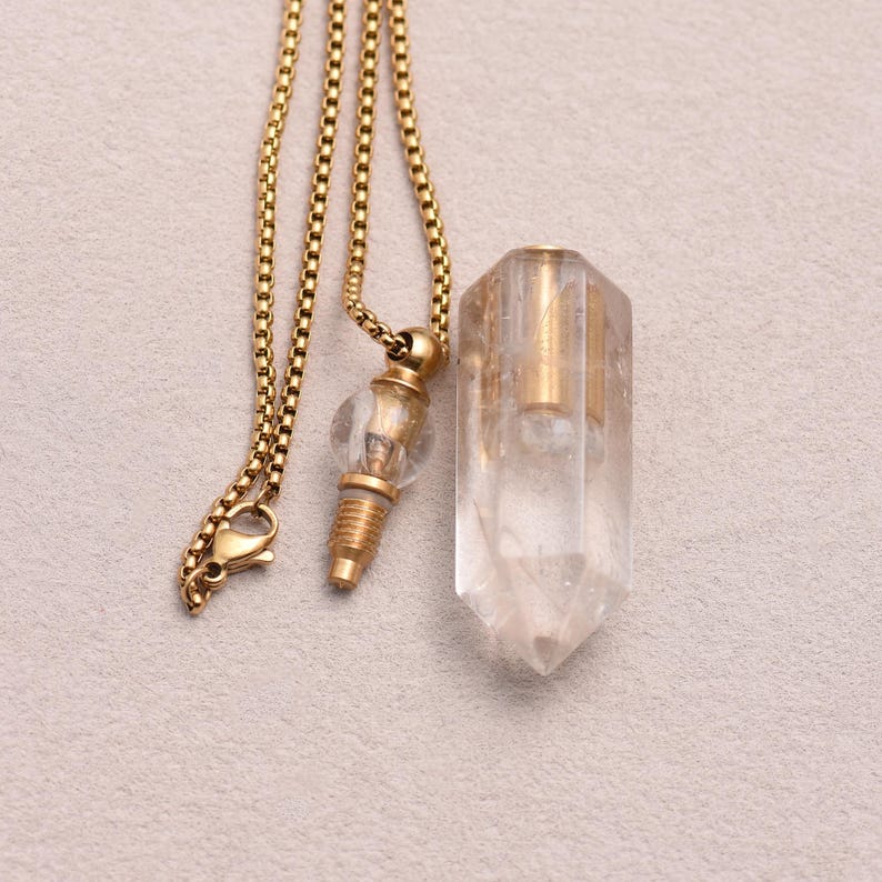 May include: A gold-colored chain necklace with two pendants. One pendant is a clear, faceted crystal. The other pendant is a small, clear glass bulb with gold-colored metal accents. The necklace is displayed on a light gray surface.