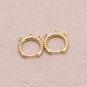 May include: Two gold-colored hoop earrings with a twisted rope design. Each earring has a small loop at the top for attachment. The earrings are displayed on a light pink surface.