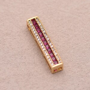 May include: A rectangular gold-toned pendant with a central row of square, deep red gemstones, flanked by rows of smaller, clear gemstones. The pendant is set against a neutral background, highlighting its intricate design and color contrast.