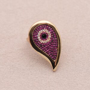 May include: Gold-coloured ring with a teardrop-shaped design. The teardrop is filled with small, vibrant magenta stones, surrounding a central eye-like detail with a black centre and white accents. The ring has a polished finish.