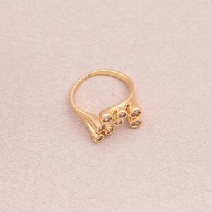 May include: A gold-colored ring with a unique design. The ring features multiple small, round, clear stones set in a flowing, organic shape. The ring is set against a light pink background, highlighting its intricate details and elegant style.