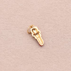 May include: A gold-toned metal charm shaped like a lipstick tube. The charm features a small circular jump ring at the top for attachment, a textured band around the center, and a smooth, elongated base with a red-colored tip at the bottom.