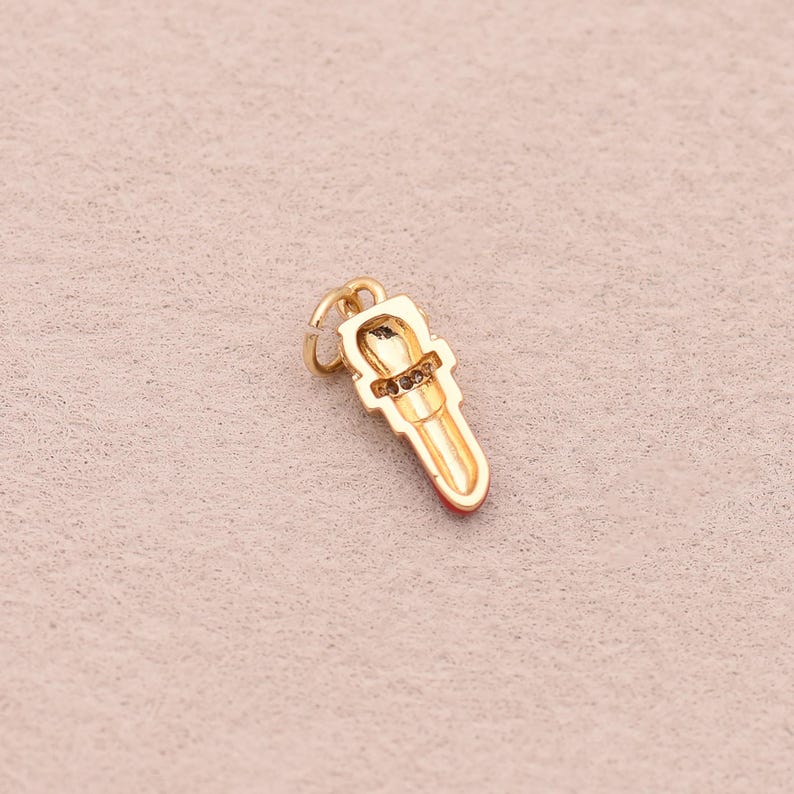 May include: A gold-toned metal charm shaped like a lipstick tube. The charm features a small circular jump ring at the top for attachment, a textured band around the center, and a smooth, elongated base with a red-colored tip at the bottom.