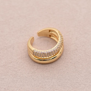 May include: A gold-colored ear cuff with a double-band design. The top band is encrusted with small, clear gemstones. The cuff has an open design, allowing it to be easily slipped onto the ear. The jewelry piece is set against a neutral background.