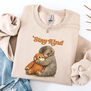 May include: Sand-colored sweatshirt with the text "Stay Kind" in orange script. The sweatshirt features an illustration of two hugging monkeys. The sweatshirt is folded on top of a pair of blue jeans.