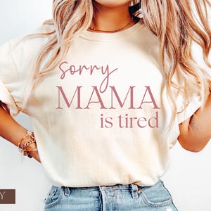 May include: Ivory-colored t-shirt with the text "Sorry Mama is tired" in a mauve color. The shirt is a crew neck style and has rolled-up sleeves. The word "IVORY" is printed at the bottom of the image.
