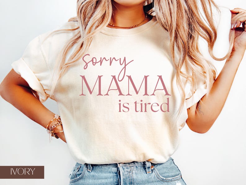 May include: Ivory-colored t-shirt with the text "Sorry Mama is tired" in a mauve color. The shirt is a crew neck style and has rolled-up sleeves. The word "IVORY" is printed at the bottom of the image.