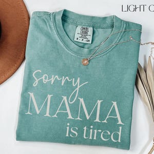 May include: Light green t-shirt with the text "Sorry Mama is tired" in white script. A gold necklace with a pendant rests on the shirt. A brown hat and dried plant are also in the image.