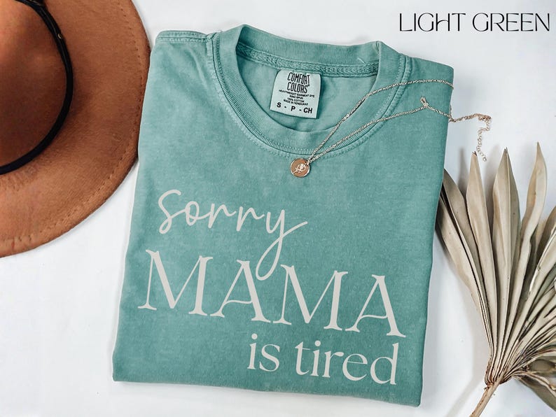 May include: Light green t-shirt with the text "Sorry Mama is tired" in white script. A gold necklace with a pendant rests on the shirt. A brown hat and dried plant are also in the image.