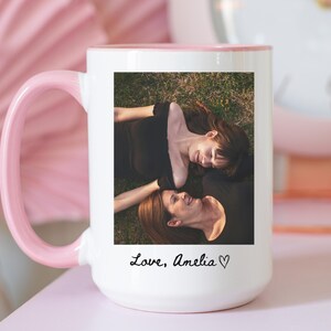 May include: White ceramic mug with a pink handle and interior. The mug features a photograph of two people lying on grass, with the text "Love, Amelia" in cursive. A pink clock is visible in the background.