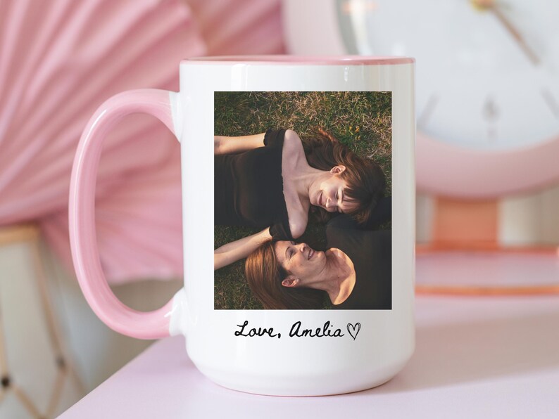 May include: White ceramic mug with a pink handle and interior. The mug features a photograph of two people lying on grass, with the text "Love, Amelia" in cursive. A pink clock is visible in the background.