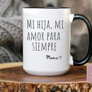 May include: White ceramic mug with a black rim and handle. The mug features the text "MI HIJA, MI AMOR PARA SIEMPRE" in black, with "Mama" and a heart symbol below. The mug is on a wooden surface.