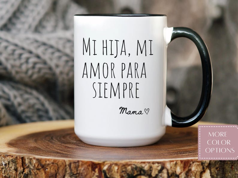 May include: White ceramic mug with a black rim and handle. The mug features the text "MI HIJA, MI AMOR PARA SIEMPRE" in black, with "Mama" and a heart symbol below. The mug is on a wooden surface.